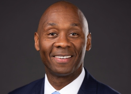 Photo of clean shaven man wearing dark blue suit and light blue tie smiling at camera