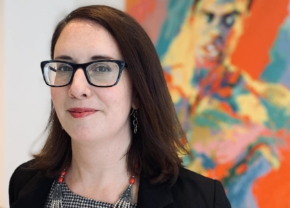 Woman with short brown hair wearing gray top, black jacket, a red necklace and black glasses smiling at camera in front of Muhammad Ali painting