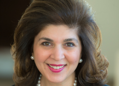 Woman with brown hair and brown eyes wearing black top and pearl necklace smiling at camera