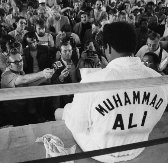 Muhammad Ali sits on the side of the ring and faces a swarm of reporters holding up microphones