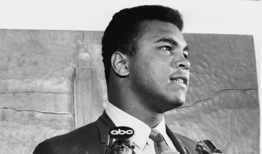 Black and white photo of Muhammad Ali in suit standing in front of microphones