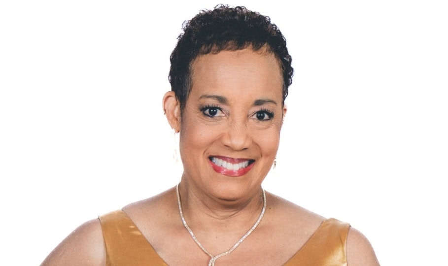 Photo of woman with short brown hair wearing necklace and gold dress smiles at camera