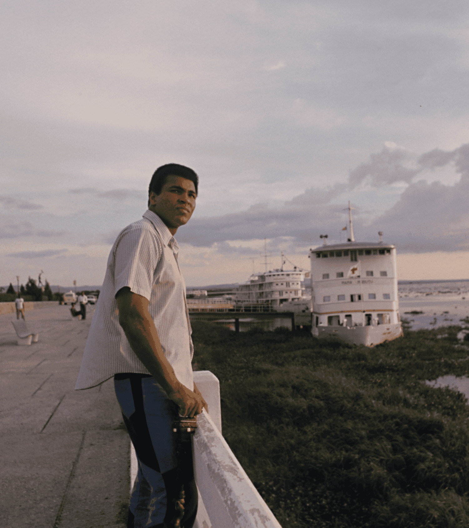 Muhammad Ali standing by the water's edge with a boat in the background