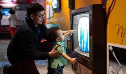 Photo of man with young boy looking at museum exhibit showcasing a television screen with Muhammad Ali