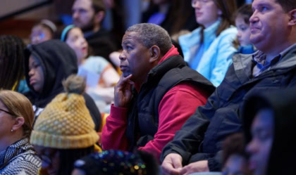 Photo of group in an auditorium with a man in the center wearing a red jacket listening intently