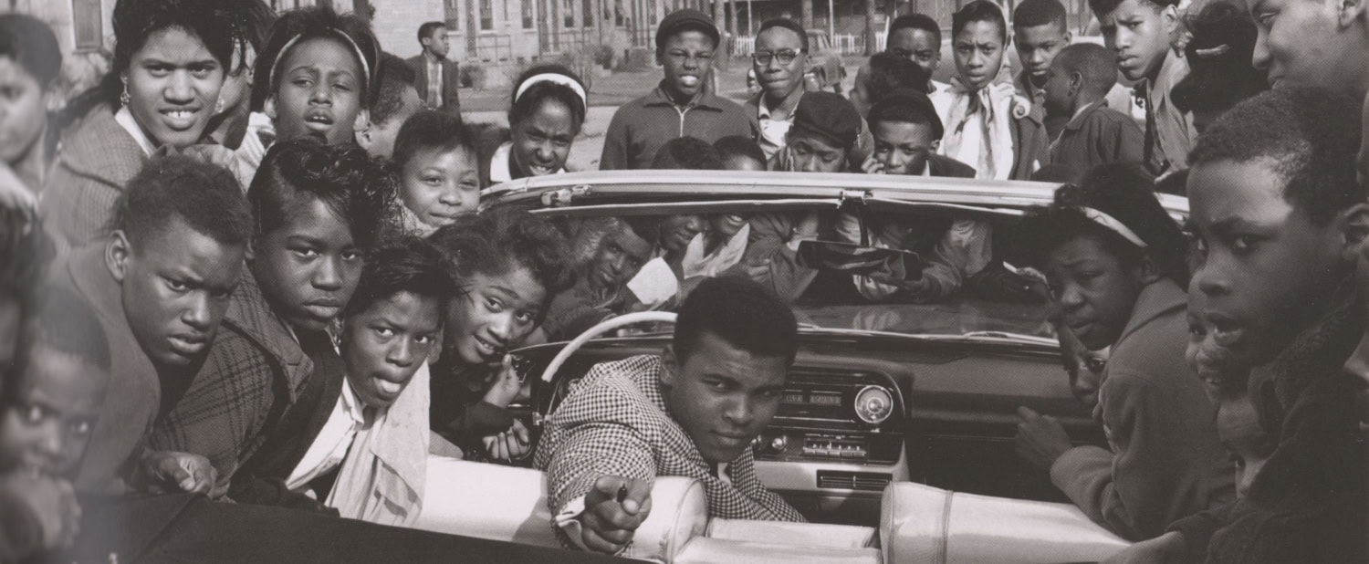 Muhammad Ali sits in car pointing backward, surrounded by a crowd