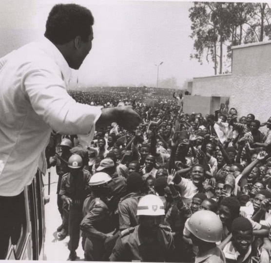 Muhammad Ali looks out a car window on to a large crowd who is cheering and raising hands