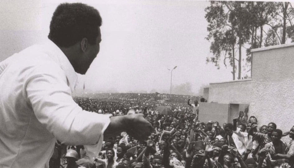 Muhammad Ali looks out a car window on to a large crowd who is cheering and raising hands