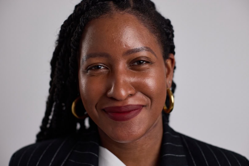 Woman with braided hair wearing gold earrings and black jacket smiles at camera