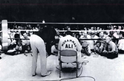 Black and white photo of Muhammad Ali taken from behind in boxing ring wearing boxing robe