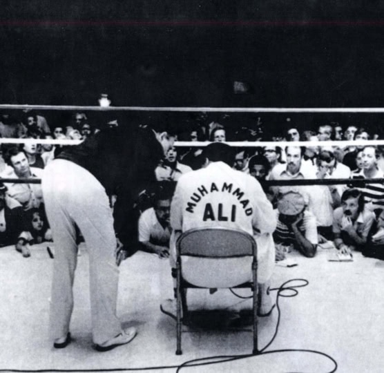 Black and white photo of Muhammad Ali taken from behind in boxing ring wearing boxing robe