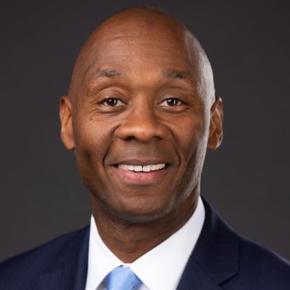 Photo of clean shaven man wearing dark blue suit and light blue tie smiling at camera