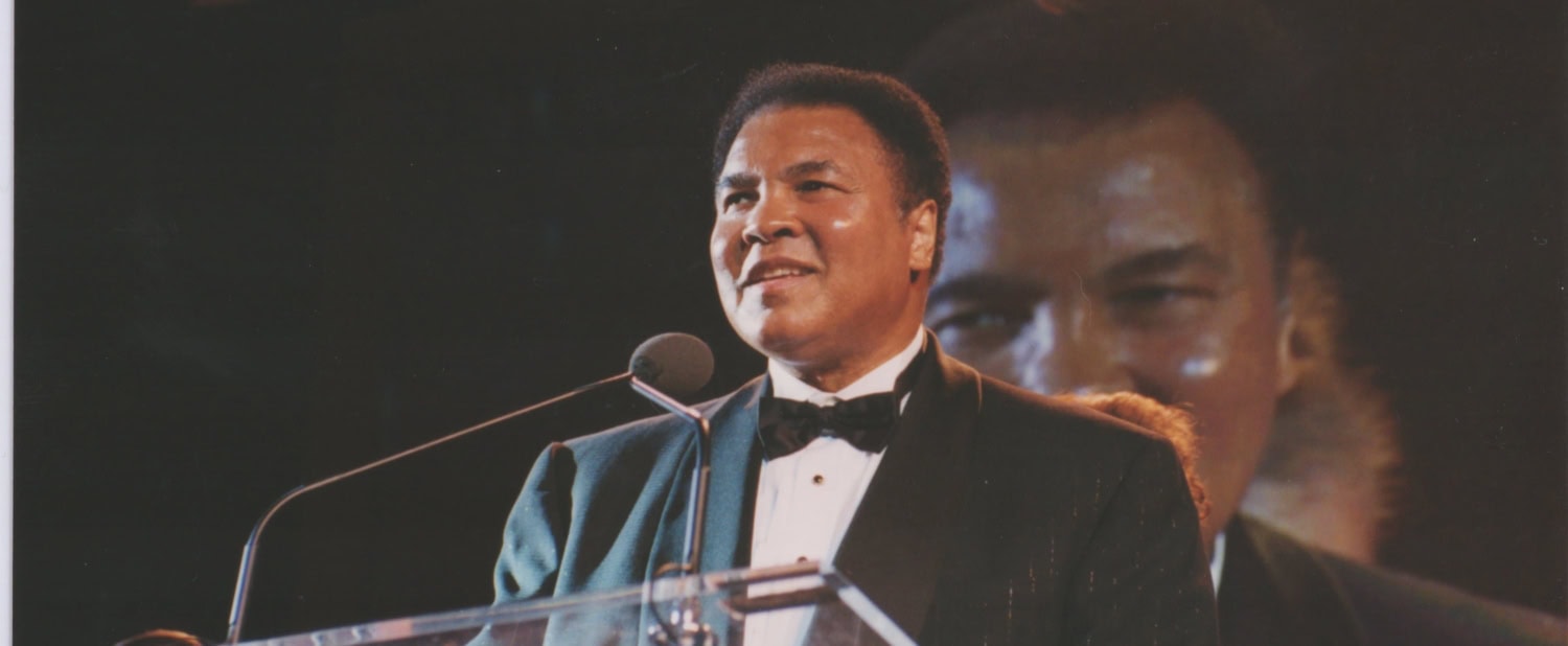 Muhammad Ali stands at a clear lectern in a tuxedo smiling