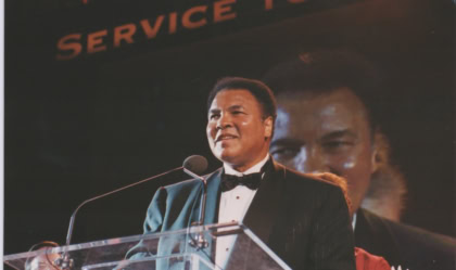 Muhammad Ali stands at a clear lectern in a tuxedo smiling