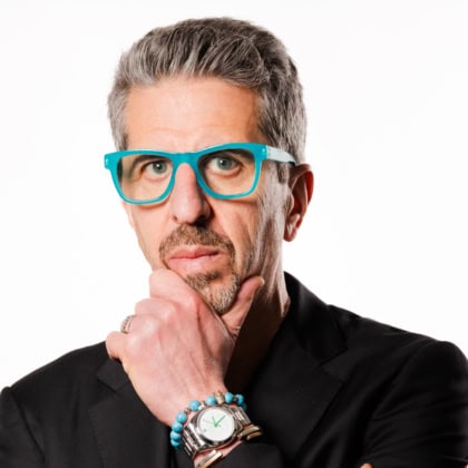 Man with short gray hair wearing blue glasses and dark suit poses with hand on chin