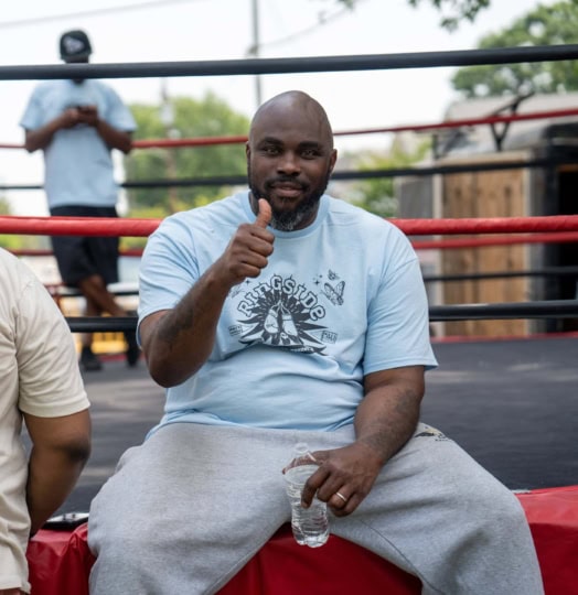 A man sits on the side of a boxing ring and gives a thumbs up