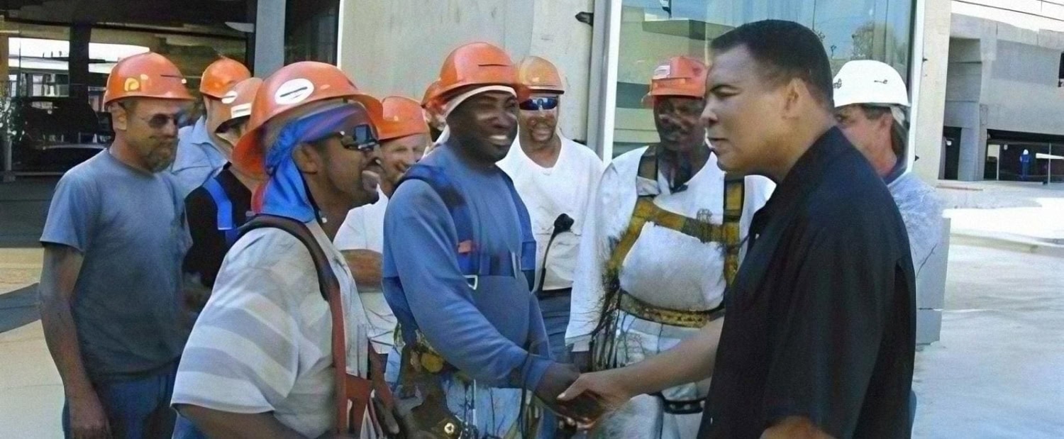 Muhammad Ali shakes hands with a man in a hard hat among a group of construction workers