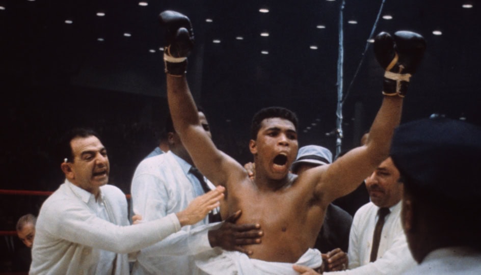 Muhammad Ali in the ring, raises his fists in victory as his corner crew surrounds him