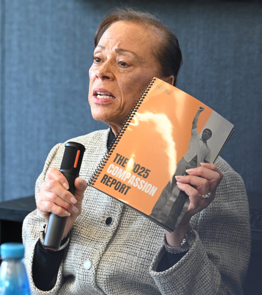 Lonnie Ali wearing light gray sweater holds up orange booklet while speaking into microphone