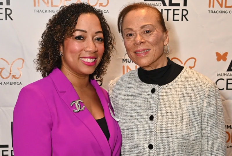 Photo of two women, one wearing pink and one wearing gray, pose for photo on Ali Index backdrop