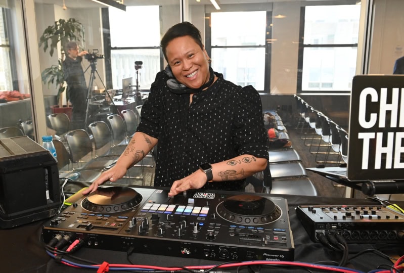 Woman behind DJ equipment with hands on turntables smiling at camera