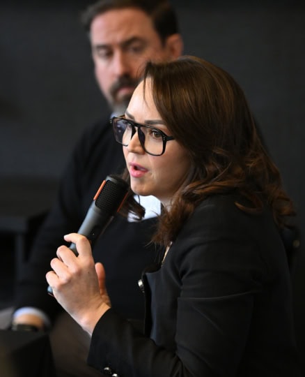 Woman with long brown hair wearing glasses speaks in microphone with man listening from behind