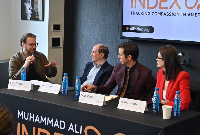 Four people sitting at table, two men and a woman are listening to man speaking into microphone