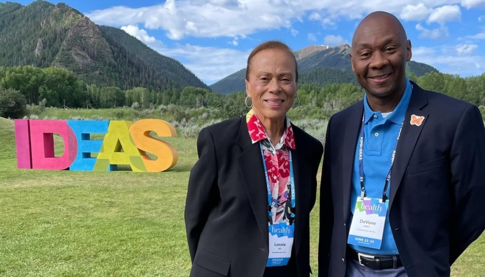 Lonnie Ali and DeVone Holt in formal wear standing outside and smiling at camera in front of small mountain range and 3D sculpture of the word 'IDEAS' in multiple colors