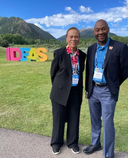 Lonnie Ali and DeVone Holt in formal wear standing outside and smiling at camera in front of small mountain range and 3D sculpture of the word 'IDEAS' in multiple colors