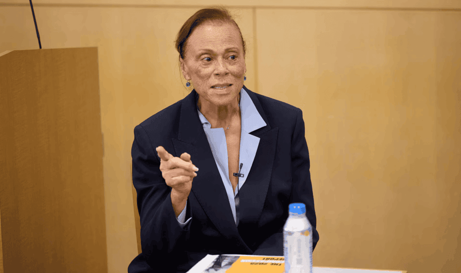 Lonnie Ali wearing dark blue blazer pointing off camera and speaking at a table