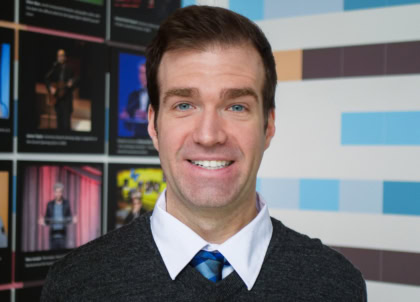 Headshot of man with short hair and blue eyes wearing gray seater and blue tie smiling at camera