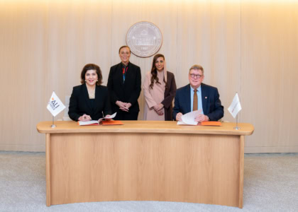 Four people sitting at wooden table and smiling at camera alongside flags with Muhammad Ali Center and American University of Sharjah logos