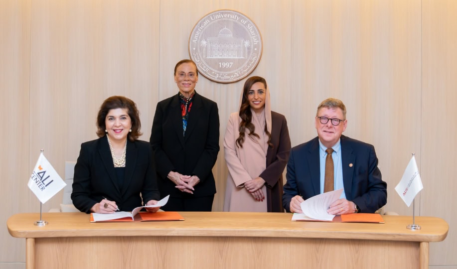 Four people sitting at wooden table and smiling at camera alongside flags with Muhammad Ali Center and American University of Sharjah logos
