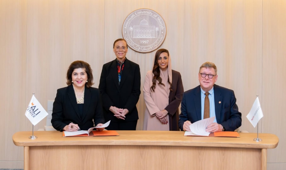 Four people sitting at wooden table and smiling at camera alongside flags with Muhammad Ali Center and American University of Sharjah logos