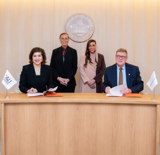 Four people sitting at wooden table and smiling at camera alongside flags with Muhammad Ali Center and American University of Sharjah logos